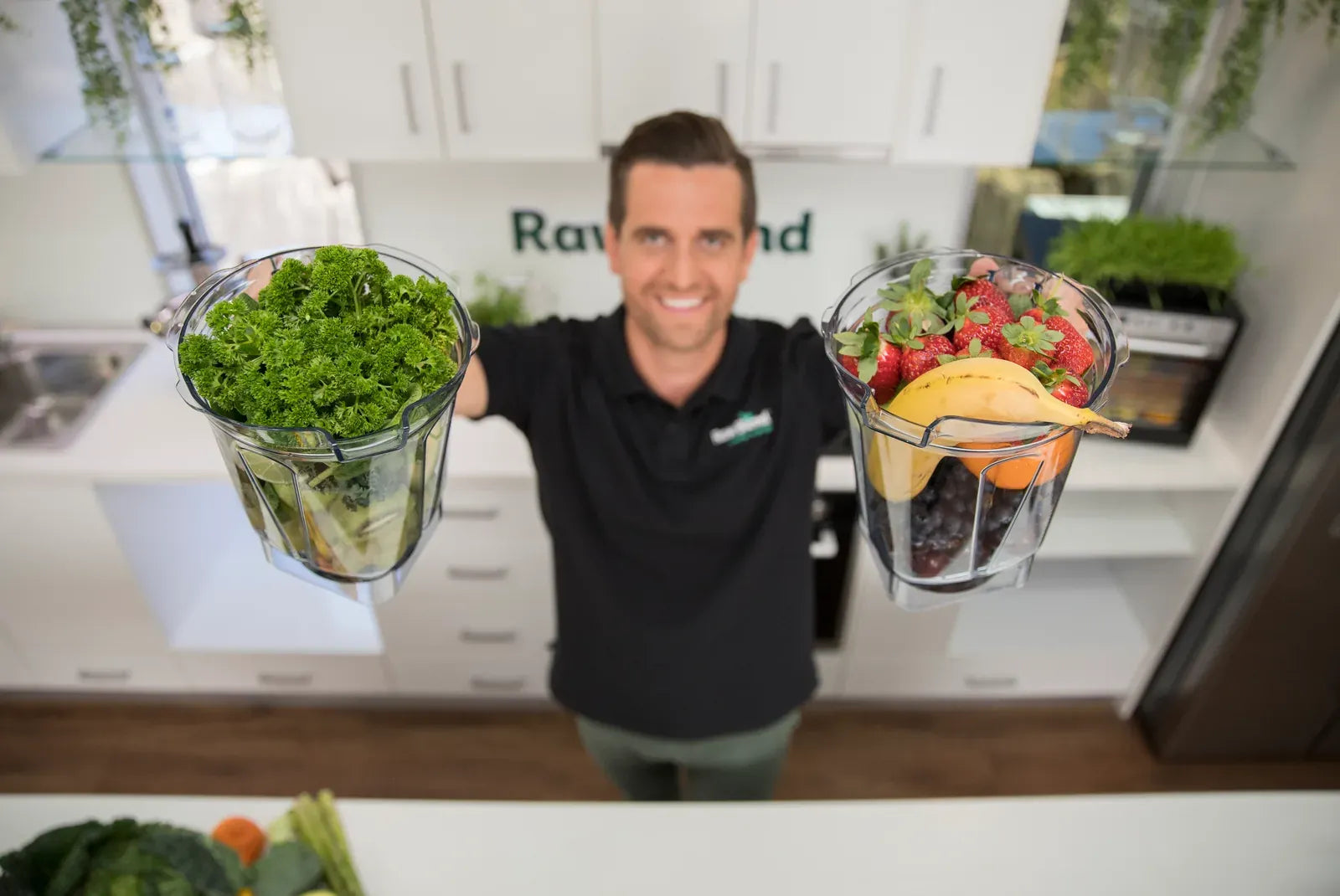 Tommy holding up two Vitamix jugs full of fresh greens and berries