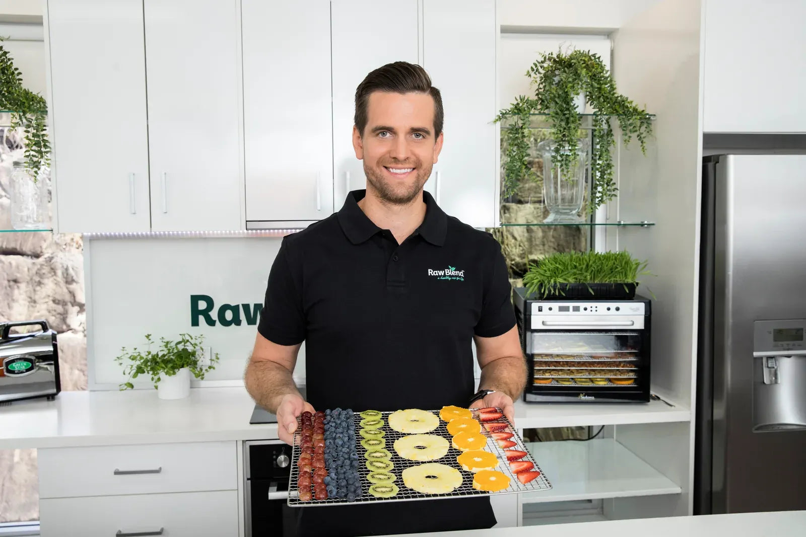 Tommy from Raw Blend smiling in the Raw Blend kitchen, holding a tray of colourful dried fruit, with dehydrators behind him