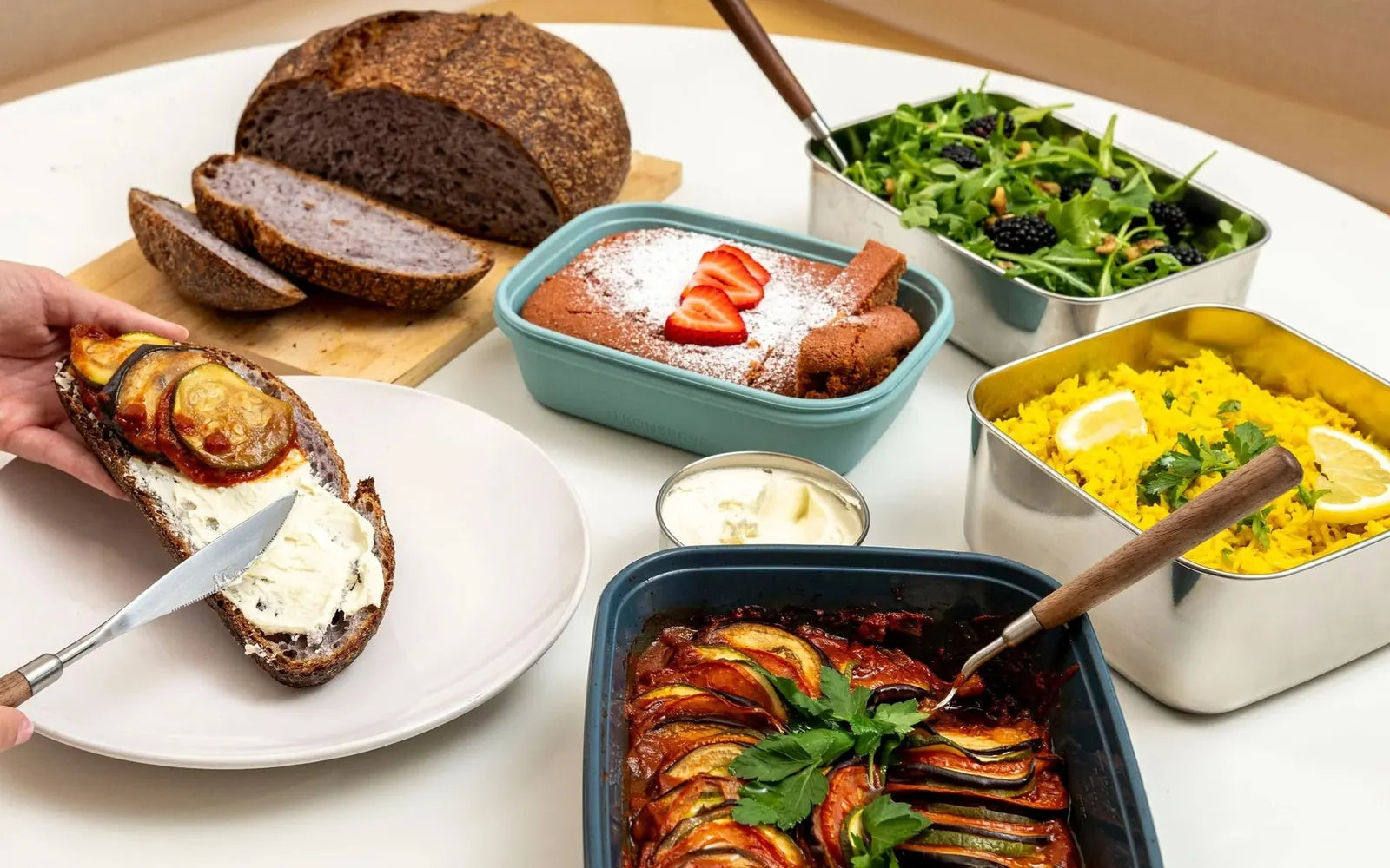 A shared lunch table with U-Konserve stainless steel containers of rocket salad and lemon rice, a baked vegetable dish and fresh sourdough