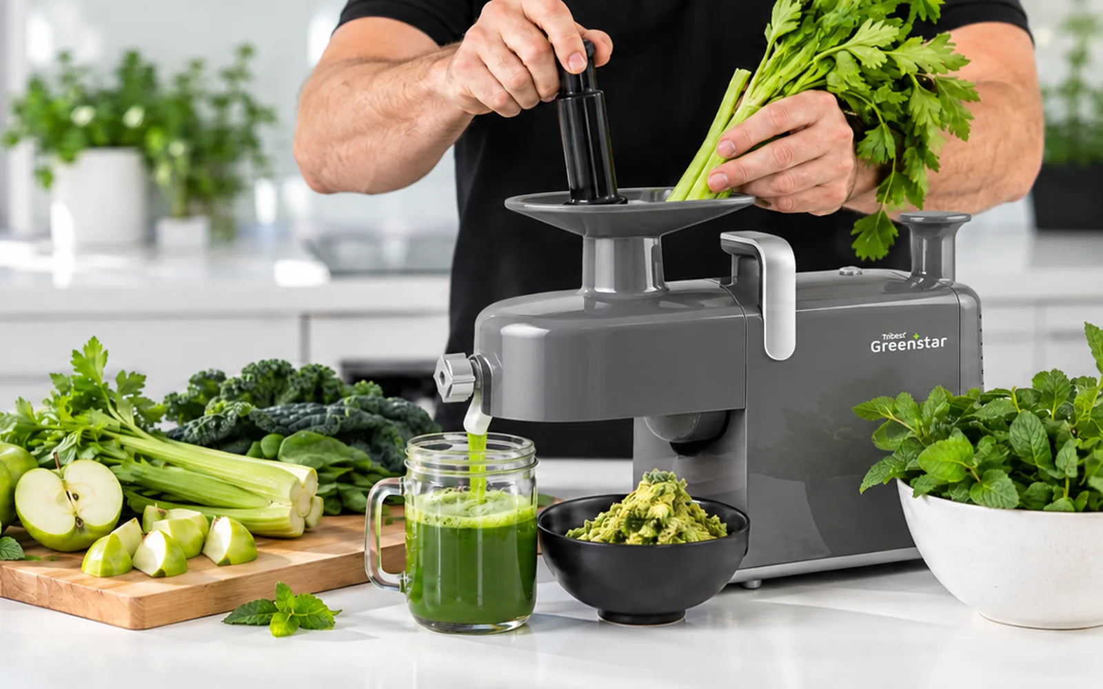 Feeding a stick of celery into a stainless steel Greenstar twin gear juicer, with sliced green apples and fresh herbs on the bench